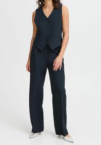 Gilet bleu marine ajusté avec un décolleté en V, quatre boutons noirs, accompagné d'un pantalon ample assorti et de talons argentés à bout pointu.