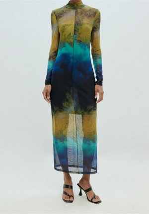 Długa sukienka z długim rękawem, sięgająca do kostek, z wysokim dekoltem, ozdobiona abstrakcyjnymi wzorami w kolorach żółtym, zielonym, niebieskim i czarnym, noszona z czarnymi sandałami na obcasie.