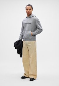 Mladý muž v béžových širokých kalhotách, šedé mikině Calvin Klein, držící černou bundu, stojící proti čistému bílému pozadí.