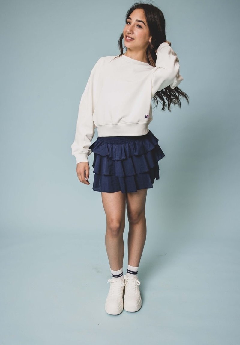 Witte sweatshirt met ribgebreide boorden, in combinatie met een marineblauwe rok met ruffles. Gemaakt met witte sneakers en gestreepte sokken. Lichte blauwe achtergrond.