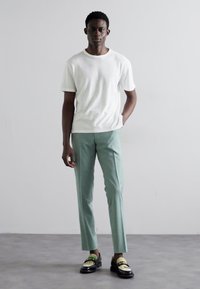 Camisa branca de manga curta, calças sob medida verdes claras, sapatos mocassins pretos com um detalhe verde. Textura suave, design minimalista, silhueta ajustada.