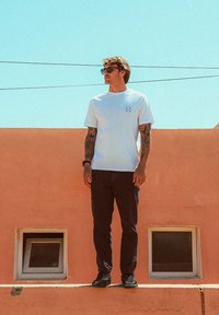 Uomo con occhiali da sole e tatuaggi si trova su un cornicione di fronte a un muro pesca con due piccole finestre sotto un cielo sereno e blu.