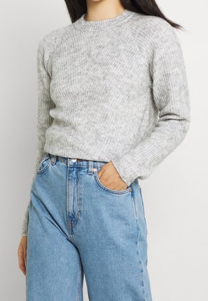 Femme portant un pull en maille côtelée gris clair et un jean denim bleu taille haute, une main dans la poche, sur fond blanc.