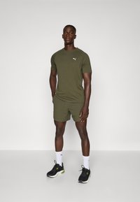 T-shirt et short de sport vert olive en tissu texturé, avec un petit logo blanc. Chaussures de sport noires avec des accents verts.