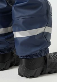 Κοντινό πλάνο ατόμου που φοράει ναυτικό αδιάβροχο παντελόνι με αντανακλαστική λωρίδα και μαύρα ανθεκτικά εξωτερικά παπούτσια σε φωτεινή επιφάνεια.