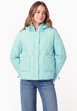 Frau trägt eine hellblaue gesteppte Kapuzenjacke mit Fronttaschen und Druckknöpfen, kombiniert mit blauen Jeans, schaut seitlich.