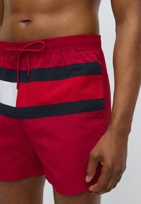 Κόκκινα μαγιό σορτς με κορδόνι στη μέση, με οριζόντιες ναυτικές και λευκές ρίγες μπροστά. Το ύφασμα φαίνεται ελαφρύ και λείο.