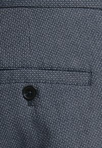 Tissu bleu foncé avec un motif texturé, présentant un seul bouton noir et un petit détail de poche.