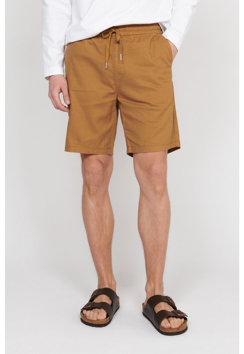 Homme portant un short beige à cordon de serrage et des sandales noires à boucle, debout devant un fond clair uni.
