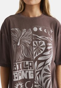 Frau trägt ein braunes T-Shirt mit weißen Blumen, Mondphasen und Sonnendesigns, mit dem Text "BILLABONG" in Blockbuchstaben.