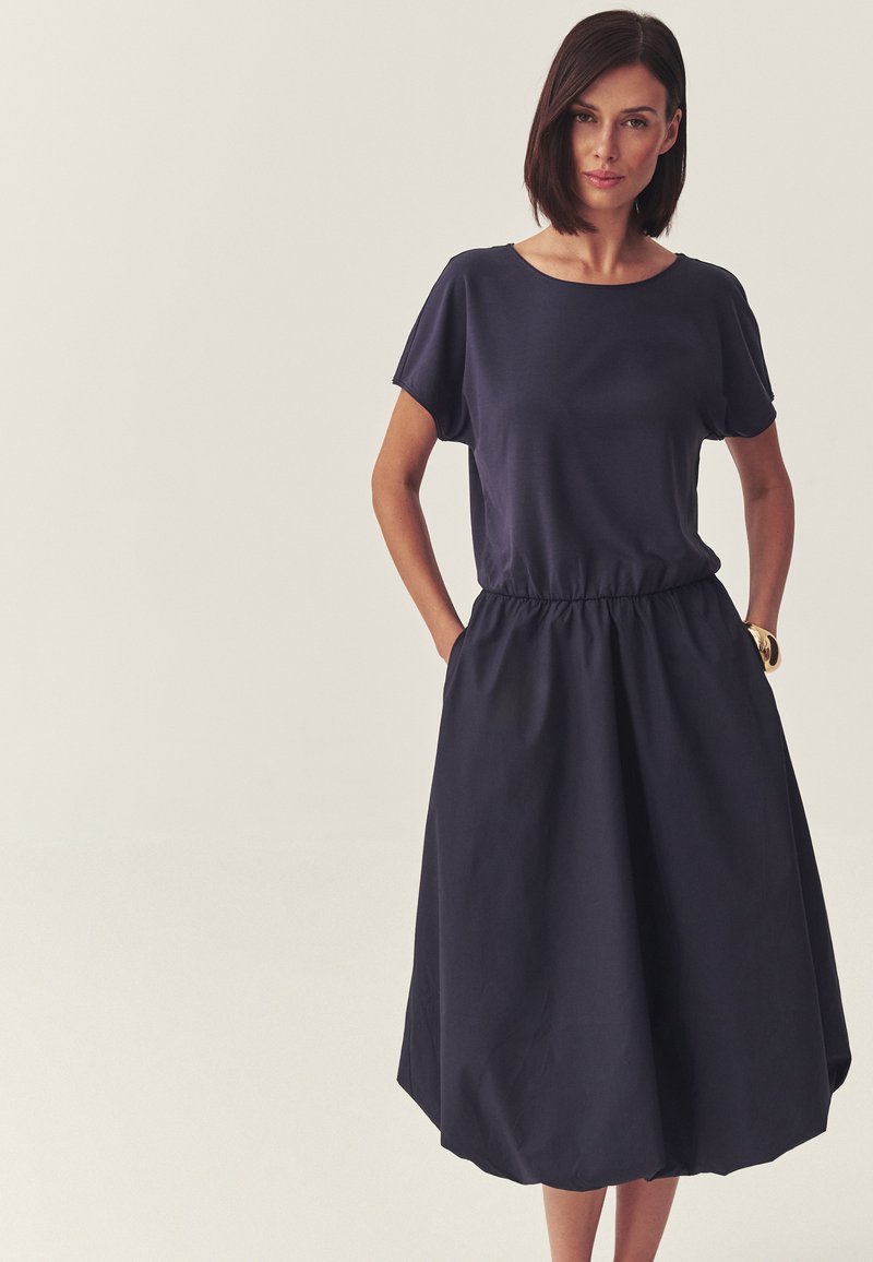Femme aux cheveux courts et foncés, portant une robe courte à manches courtes bleu marine ample avec une taille élastique et les mains dans les poches, debout devant un fond clair.