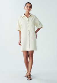 Šaty do kolen s knoflíkovou légou v světle žluté barvě, s krátkými rukávy a náprsní kapsou. Vyrobené z texturované látky s volným střihem.
