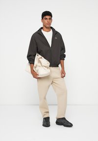 Giacca nera leggera con zip, pantaloni beige, sneaker nere e una borsa a spalla crema. Design semplice con un branding sottile.