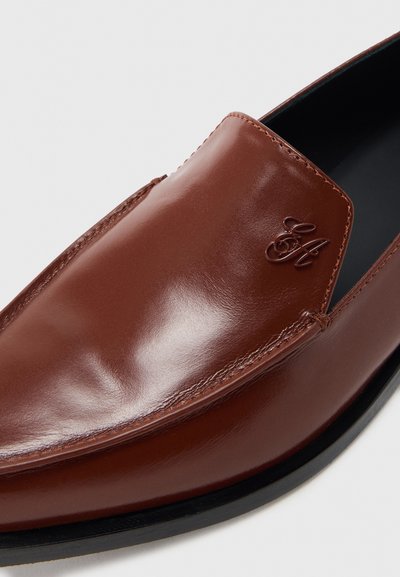 Mocasini din piele maro cu finisaj lucios, având un design cusut și un logo embossat pe lateral. Talpă neagră cu accente minimaliste.