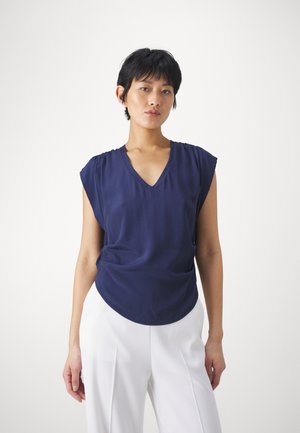 Top blu navy a maniche corte con scollatura a V e morbidi drappeggi. Tessuto liscio e orlo arrotondato, abbinato a pantaloni bianchi a vita alta.