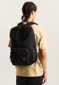 Persoon met vlechten, gekleed in een beige shirt en een zwarte Vans rugzak met voorvak en gespsluiting, met de rug naar de camera gericht tegen een effen achtergrond.