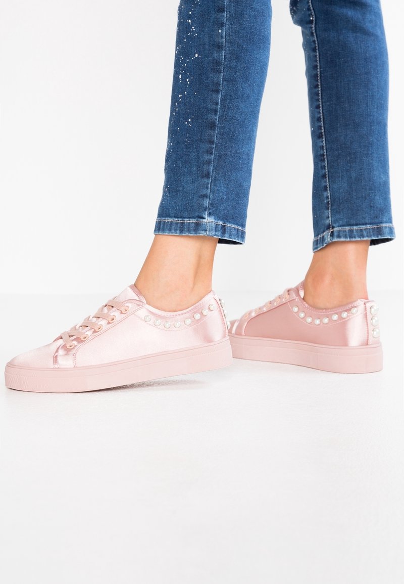 Personne portant des baskets rose clair brillantes avec des détails en perle, associées à un jean bleu avec des éclaboussures de peinture blanche sur fond blanc.