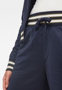 Mörkblå träningsoverall med randig midja i beige och vitt, utrustad med dragsnöre och ribbade cuffar. Slät textur och avslappnad passform.