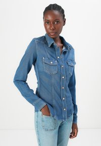 Camicia in denim blu con design aderente, colletto a punta, due tasche sul petto e accessori in rame. Maniche lunghe con polsini abbottonati.