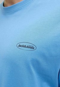 Světle modré bavlněné tričko s kulatým výstřihem a krátkými rukávy. Na hrudi má černý potisk loga "Jack & Jones Club 1990."