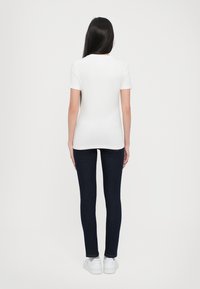 T-shirt branca de manga curta combinado com jeans skinny azul-escuro e ténis brancos, visto de costas contra um fundo simples.