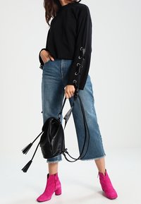 Mujer vistiendo una blusa negra de mangas largas con cordones en las mangas, jeans cortos azules, botas de tobillo rosas brillantes, sosteniendo una mochila de cuero negro.