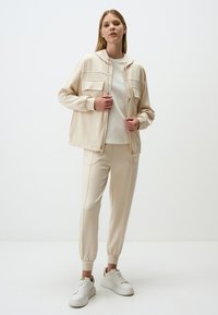 Beige loungewear set met een ritsjack met twee voorkruimelzakken en bijpassende taps toelopende broek. Het materiaal lijkt zacht met een gladde textuur.