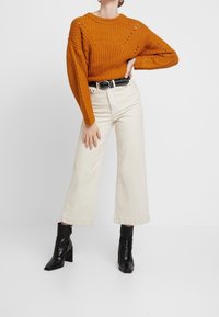 Fetziges orange gestricktes Sweatshirt mit diagonalen Mustern, hochgeschlossen, kombiniert mit weiten beigen Hosen und schwarzen Stiefeletten mit Absatz.