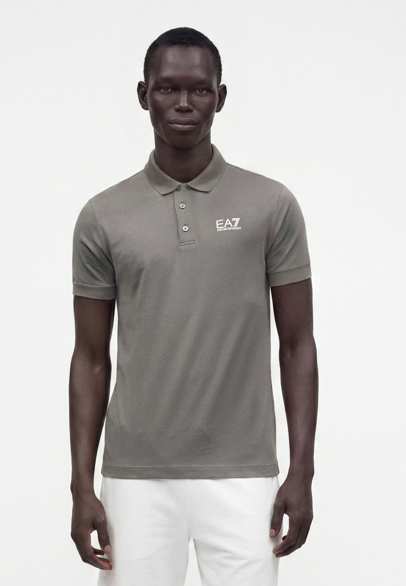 Homme portant un polo gris à manches courtes avec le logo EA7 Emporio Armani et un pantalon blanc, debout devant un fond uni.