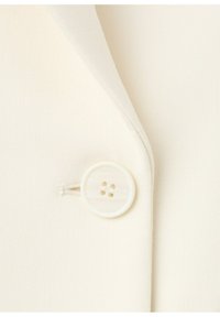 Flødefarvet stof med en enkelt rund knap med fire huller, fastgjort nær en foldet kant, der fremhæver beklædningsdetalje og tekstur.