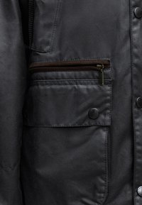 Gros plan d'une poche de veste noire avec un compartiment zippé marron au-dessus d'une poche à rabat boutonnée, montrant des coutures et des boutons détaillés.