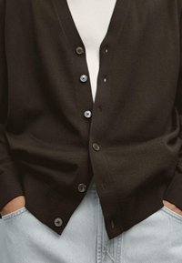Brun stickad cardigan med V-ringning och sex framsidor knappar. Har ribbade ärmslut och fåll, bärs över en vit skjorta och kombineras med ljus jeans.