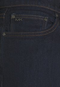 Zbliżenie na ciemnoniebieskie dżinsy z kieszenią z srebrnym logo MK oraz kontrastującym pomarańczowym przeszyciem wzdłuż szwów.