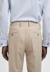 Beige tailored trousers with double pleats, side pockets, and two back pockets featuring button accents. Paired with a light blue shirt.