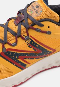 Gros plan d'une chaussure de sport jaune avec des lacets gris foncé, un graphisme rouge et noir sur le côté, et une étiquette "Fresh Foam Garde" sur la languette.