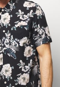 Mężczyzna w czarnej koszuli z krótkim rękawem zapinanej na guziki, ozdobionej białymi i zielonymi wzorami kwiatów i liści, prezentujący górną część torsu i ramienia.