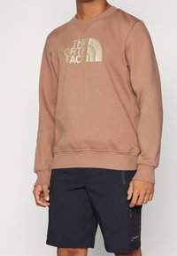 Sudadera marrón claro con cuello redondo, que presenta un gran logotipo bordado en oro. Combinada con pantalones cortos azul oscuro con bolsillos con cremallera.