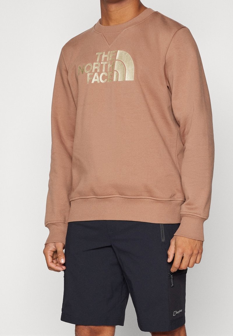 Sudadera marrón claro con cuello redondo, que presenta un gran logotipo bordado en oro. Combinada con pantalones cortos azul oscuro con bolsillos con cremallera.