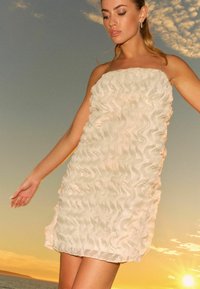 Robe mini créme à volants avec fines bretelles, présentant une texture ondulée sur l'ensemble, sur fond de coucher de soleil.