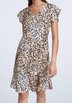 Frau trägt ein beige-schwarz gepunktetes, kurzärmliges V-Ausschnitt-Kleid mit ausgestelltem Saum vor einem schlichten hellen Hintergrund.