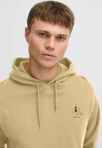 Beige hettegenser med lomme foran, snor og en brodert "Reward"-logo som viser drikker og en bok. Myk, bomullsblanding.