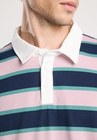 Lyhythihainen poolopaita, joka on valmistettu pehmeästä kankaasta ja jossa on horisontaaliset raidat vaaleanpunaisena, laivastonsinisenä ja tealina. Valkoinen kaulus, jossa on kolmen napin sulkeminen.