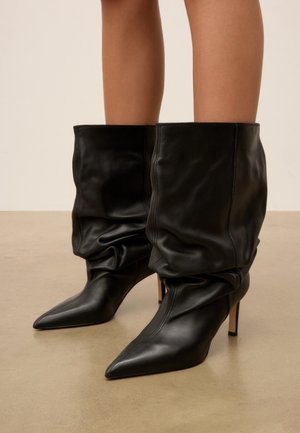 Bottes en cuir noires souples mi-mollet avec bouts pointus et talons hauts et fins, portées par une personne debout sur un sol lisse.