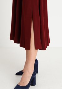 Jupe plissée cramoisie avec une fente sur le côté, fabriquée en tissu doux, assortie à des chaussures à talons hauts en suède bleu marine, avec un design de talon large.