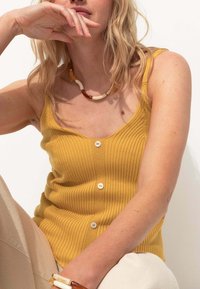 Débardeur côtelé moutarde avec un décolleté en V, comportant trois boutons blancs. Assorti à un pantalon de couleur neutre et un collier multicolore.