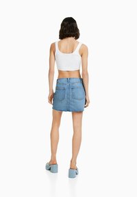 Donna in piedi di spalle, che indossa un top corto bianco, una mini gonna di jeans blu con tasche posteriori e scarpe con tacco a blocco di un blu chiaro.