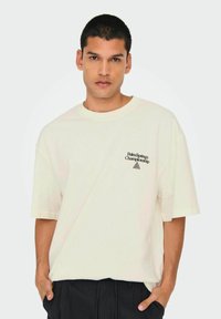 Camiseta oversized color crema de tejido suave con un logotipo en el pecho que dice "Palm Springs Championship" y un acento en forma de triángulo.