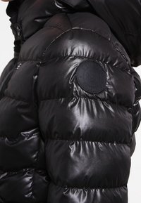 Zbliżenie na błyszczącą czarną pikowaną kurtkę puchową z okrągłą czarną naszywką logo na górnej części rękawa.