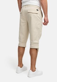 Shorts cargo beiges avec des revers, en coton, dotés de deux poches arrière et d'une petite étiquette noire. Portés avec des baskets blanches.