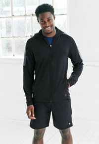 Musta urheiluhuppari vetoketjulla ja huppulla, mustat shortsit, kevyt kangas, kapea istuvuus ja vetoketjut yksityiskohtina.